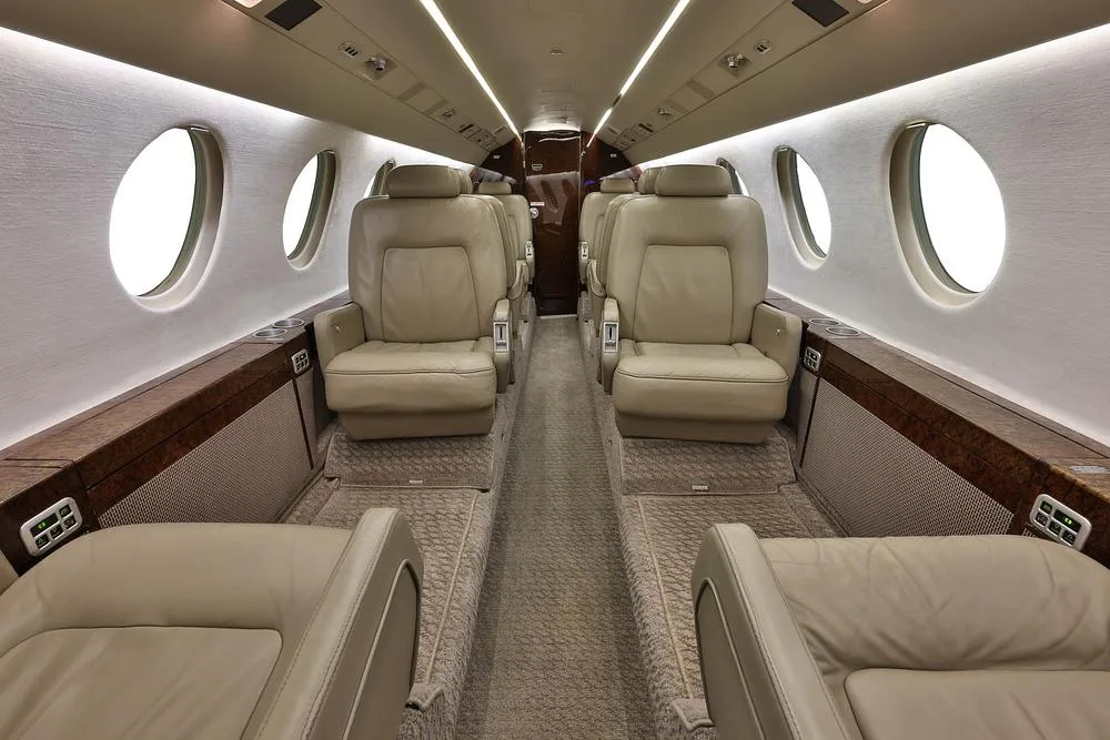 The luxurious cabin interior of a business jet with beige leather seats and wood trim.