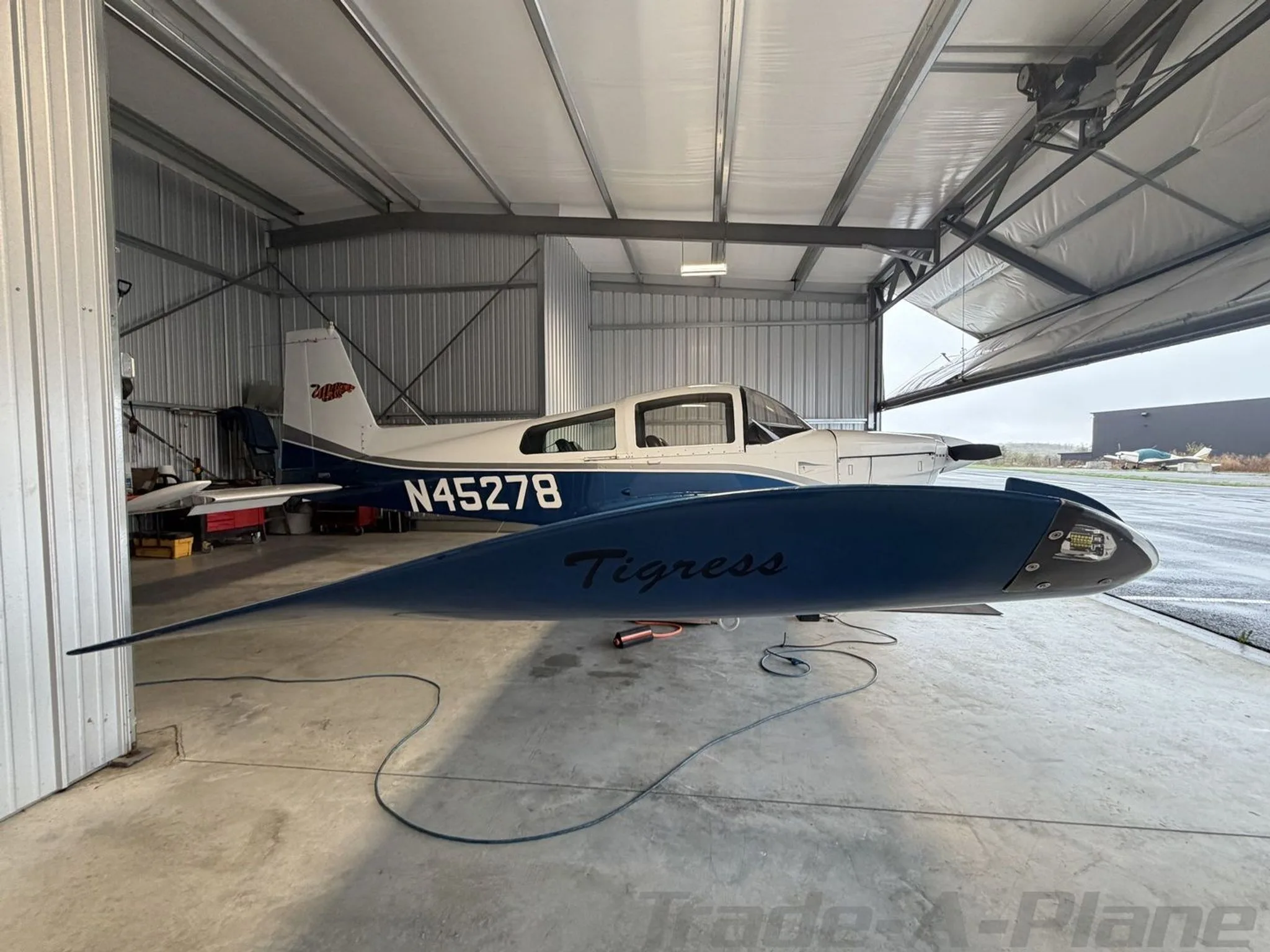 Side view of a white and blue Grumman AA-5B Tiger N45278 in a hangar, showing the 'Tigress' decal on the wing.