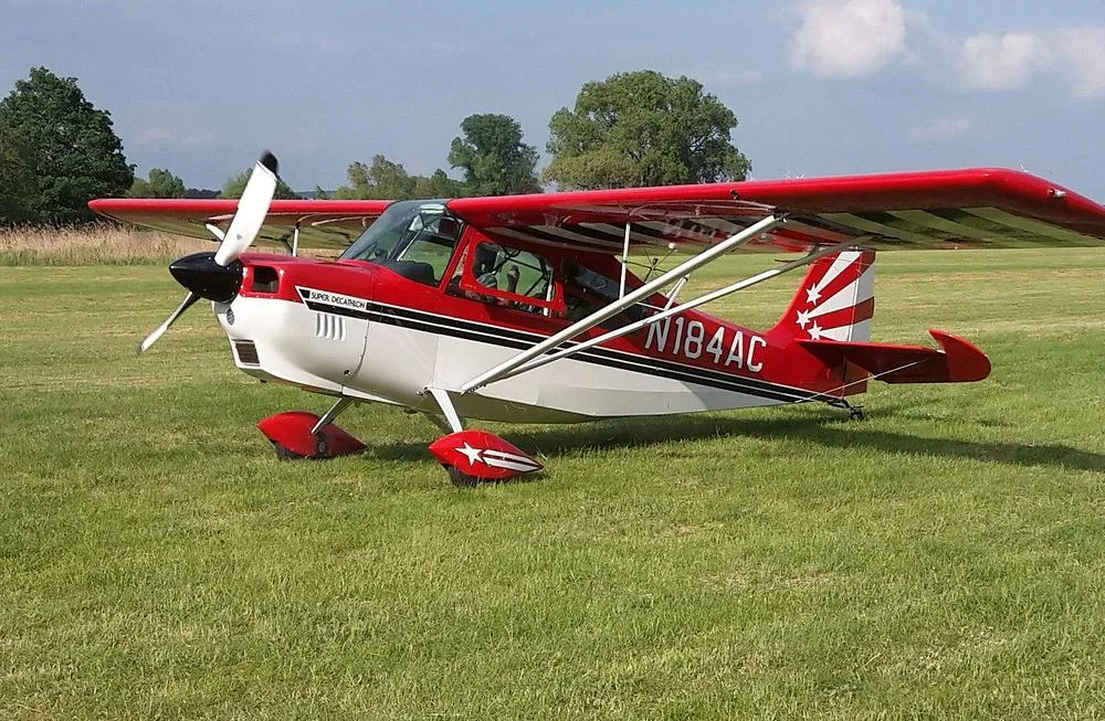 A red and white Super Decathlon aircraft (N184AC) with a striped tail is parked on a grassy field.