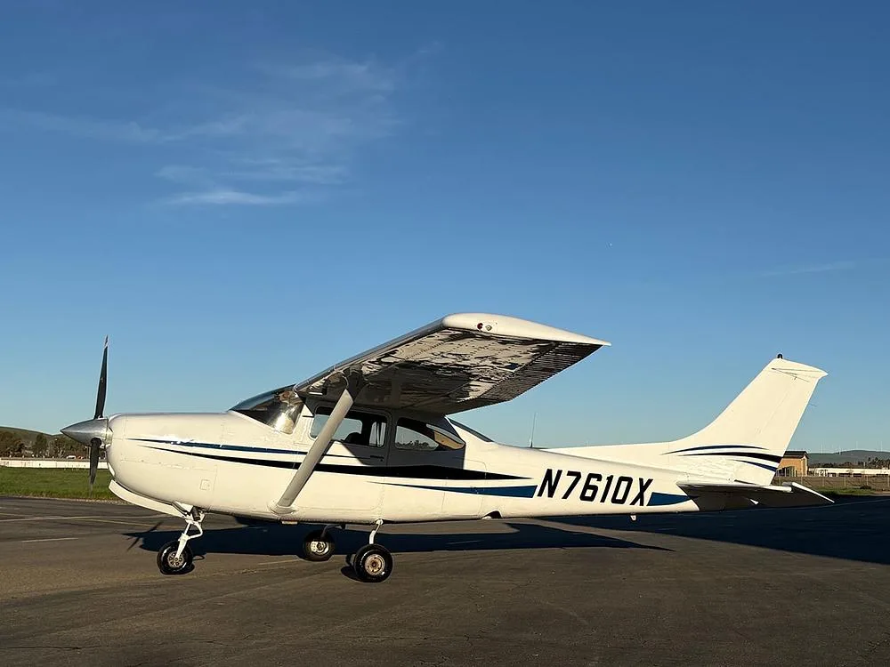 A white Cessna 172 with blue and black stripes, registration N7610X, parked on an asphalt ramp under a blue sky.