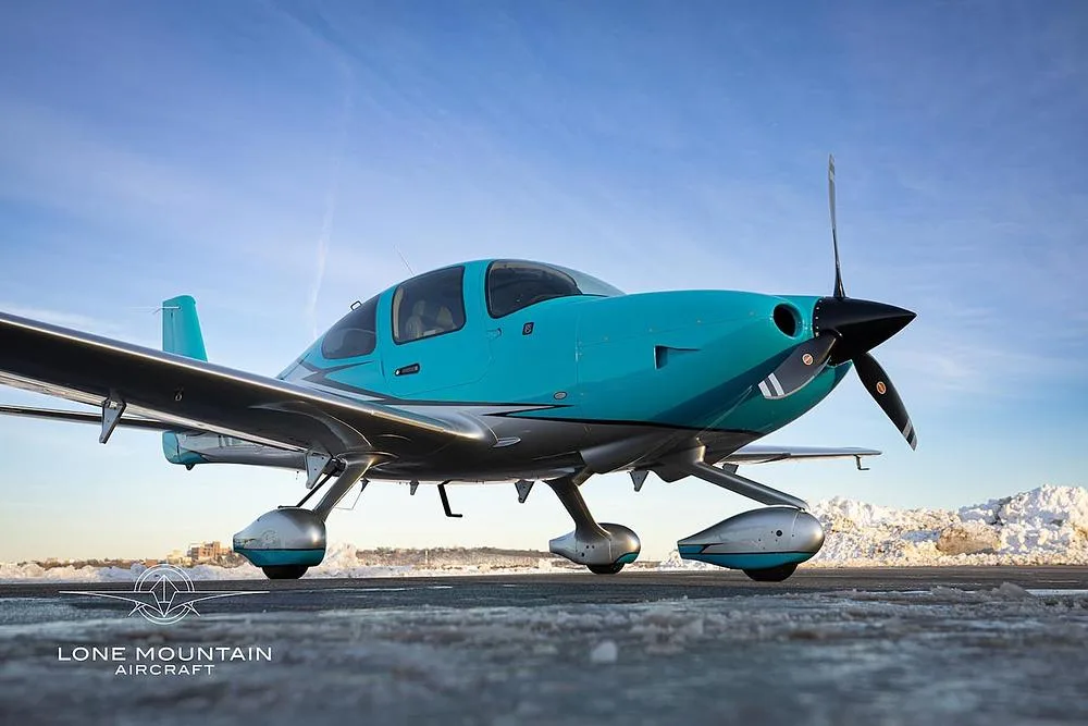 A low-angle front view of a turquoise and silver single-engine aircraft on a snowy tarmac.
