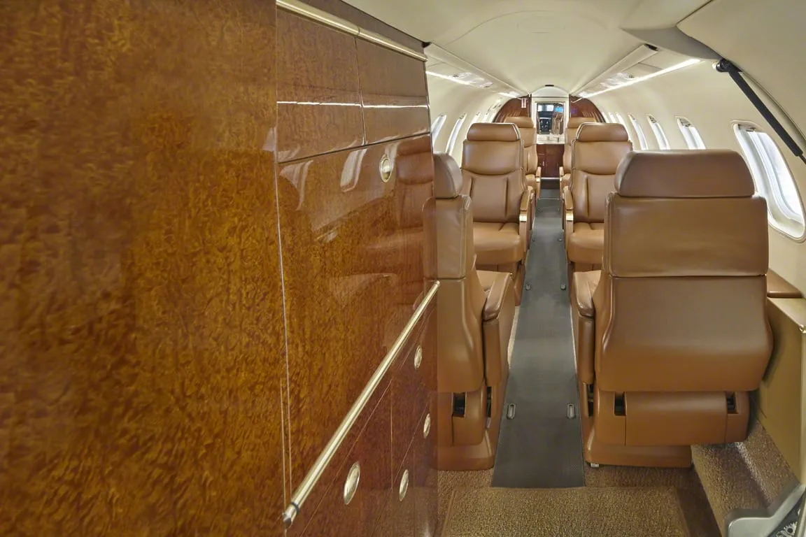 A long view of a private jet's luxurious cabin with tan leather seats and glossy wood paneling.