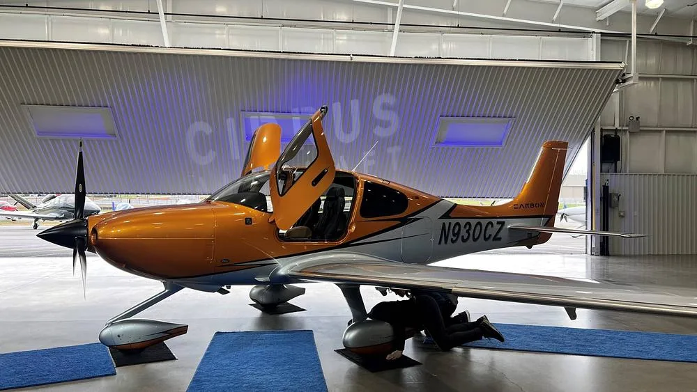 A shiny orange and silver Cirrus SR22 aircraft with its doors open, parked inside a hangar with "CIRRUS" projected on the back wall.
