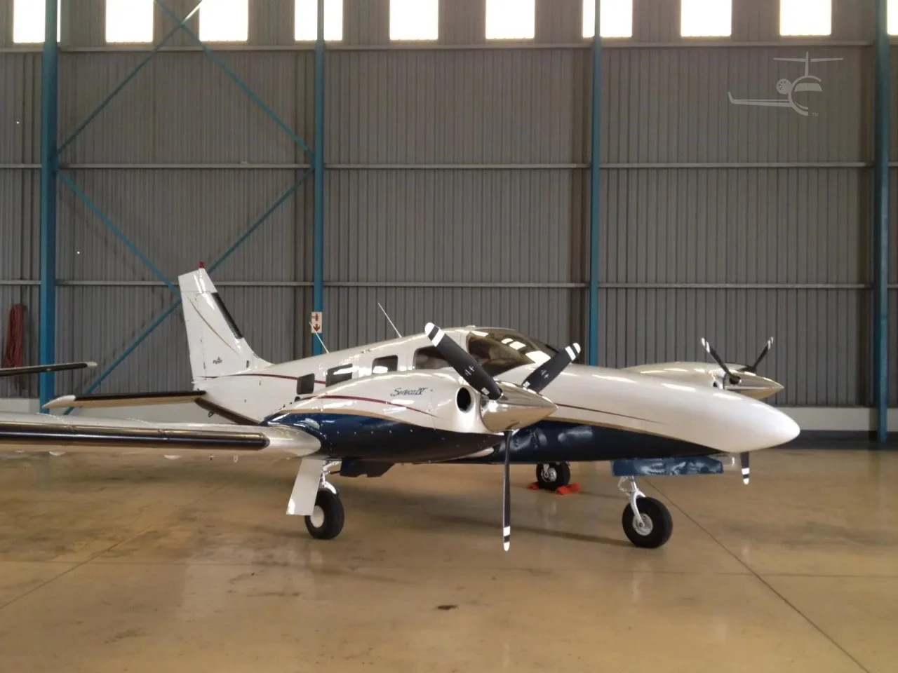 A white and blue Piper Seneca II twin-engine aircraft is parked inside a hangar.
