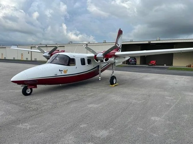 A white and red twin-engine propeller aircraft parked on an asphalt ramp with hangars in the background.