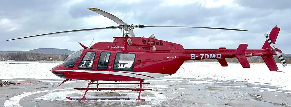 A red Bell 407GXP helicopter with white stripes, tail number B-70MD, parked on a snowy helipad under a cloudy sky.