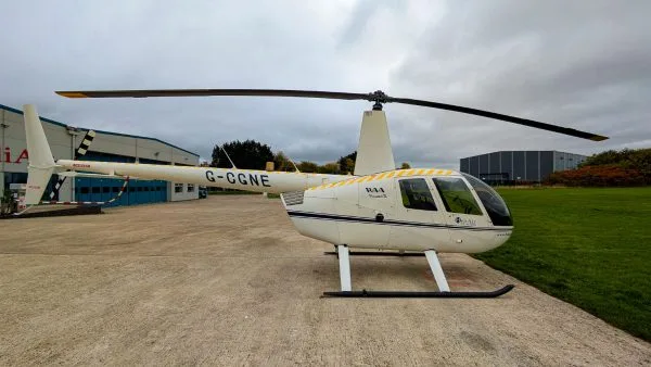 A white Robinson R44 Raven II helicopter with registration G-CGNE is parked on an airfield tarmac next to hangars.