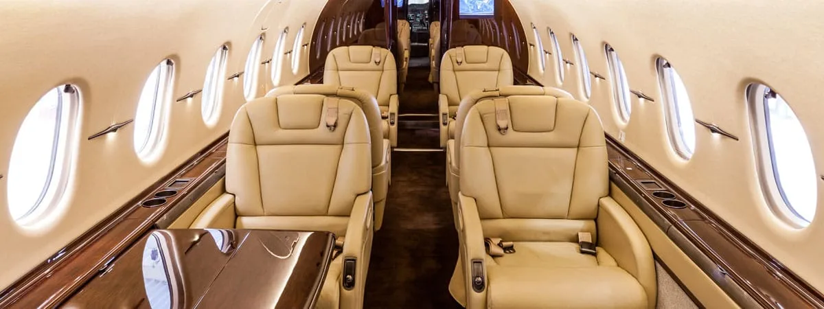 A view down the aisle of a luxurious aircraft cabin with tan leather seats and dark wood accents.