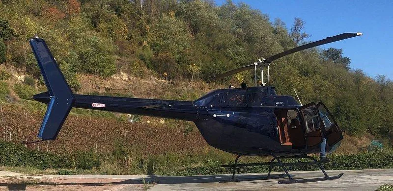 A dark blue Bell 206 helicopter is parked on a concrete pad with its door open, against a backdrop of autumn trees.