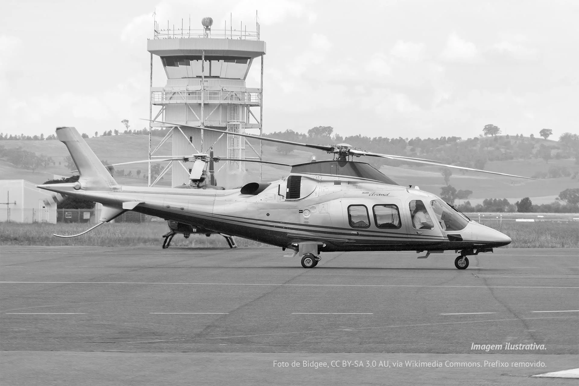 A black and white AgustaWestland AW109 Grand helicopter on an airfield with an air traffic control tower in the background.