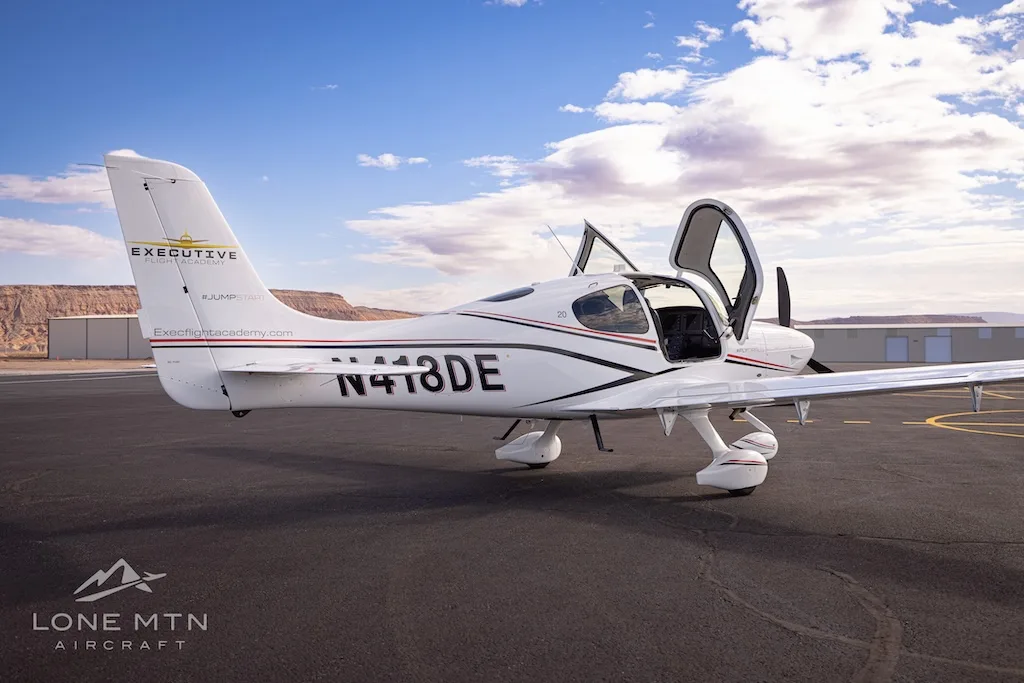A white Cirrus SR22 aircraft with black and red stripes, registration N418DE, parked on an airfield with doors open.