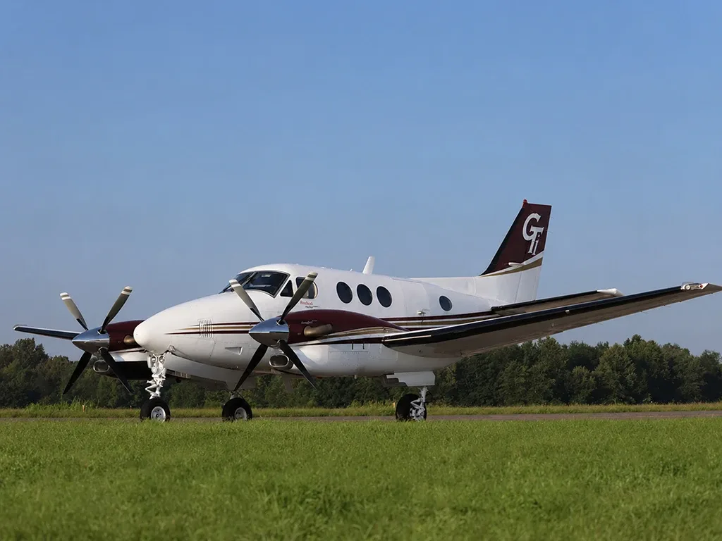 A white and maroon twin-turboprop aircraft with a 'GT' logo on its tail sits on a grassy airfield under a clear blue sky.