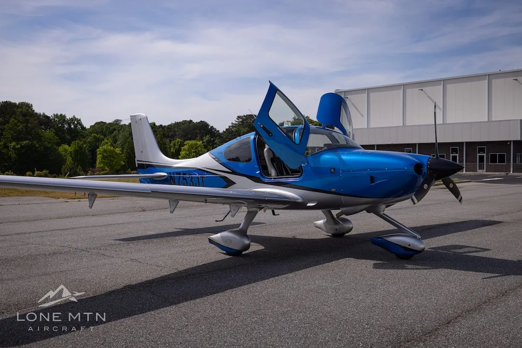 A blue and silver single-engine aircraft with open gull-wing doors is parked on an asphalt tarmac.