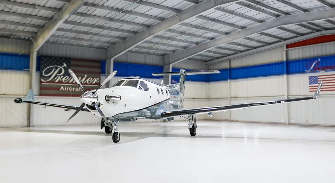 A front view of a white and grey Pilatus PC-12 turboprop aircraft, tail number N411PC, parked inside a hangar.