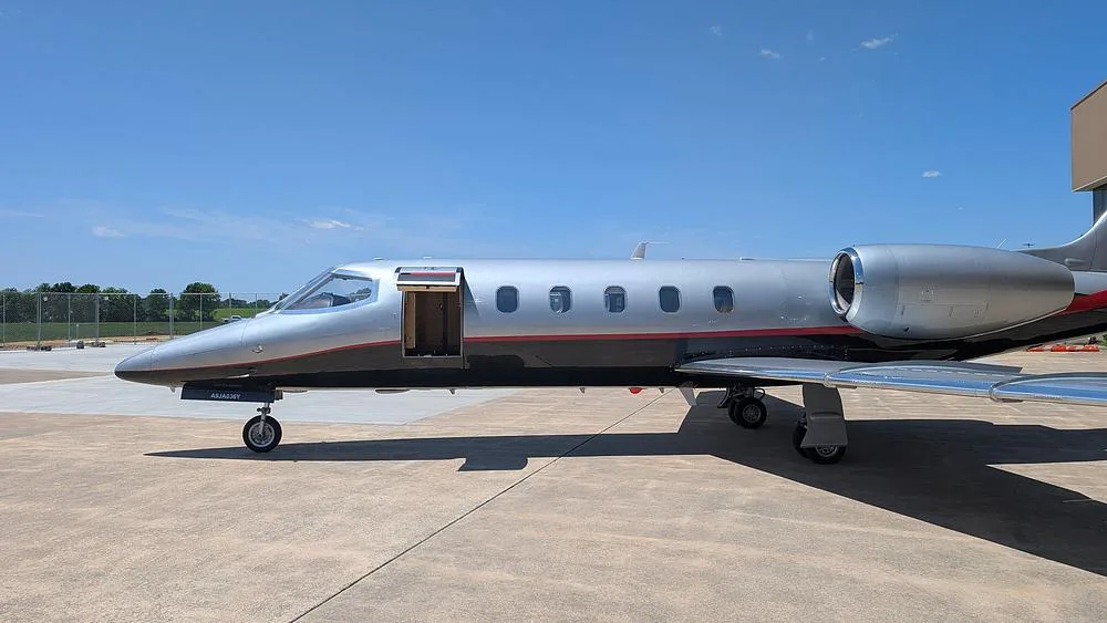 A silver and black Learjet with a red stripe, parked on a tarmac with its main cabin door open, under a blue sky.