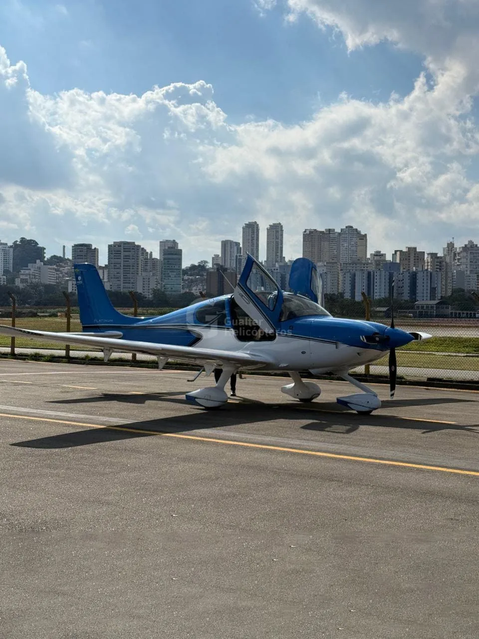 A blue and white Cirrus SR22 aircraft with its doors open, parked on an airfield with a city skyline in the background.