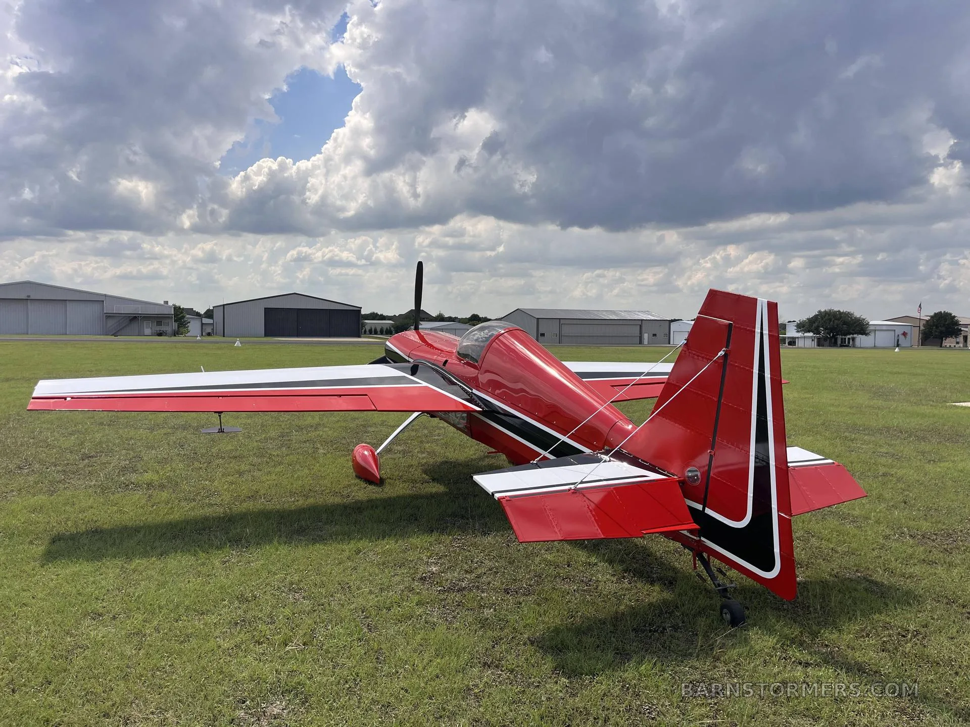 A red and white aerobatic aircraft, N300CB, parked on a grassy airfield with hangars in the background under a cloudy sky.