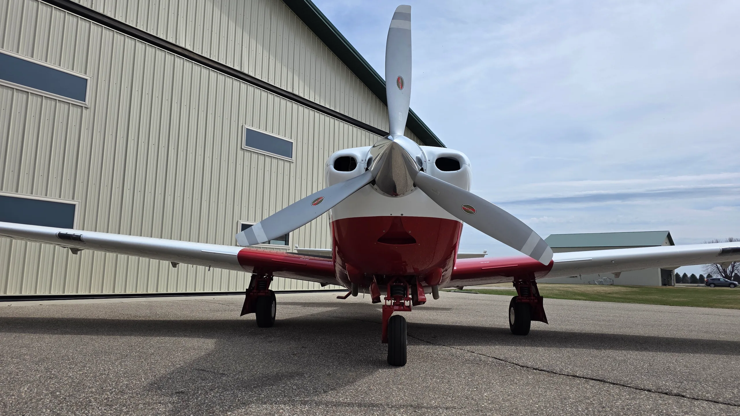 A head-on view of a red and white Mooney aircraft with a three-bladed propeller, parked on an asphalt apron.
