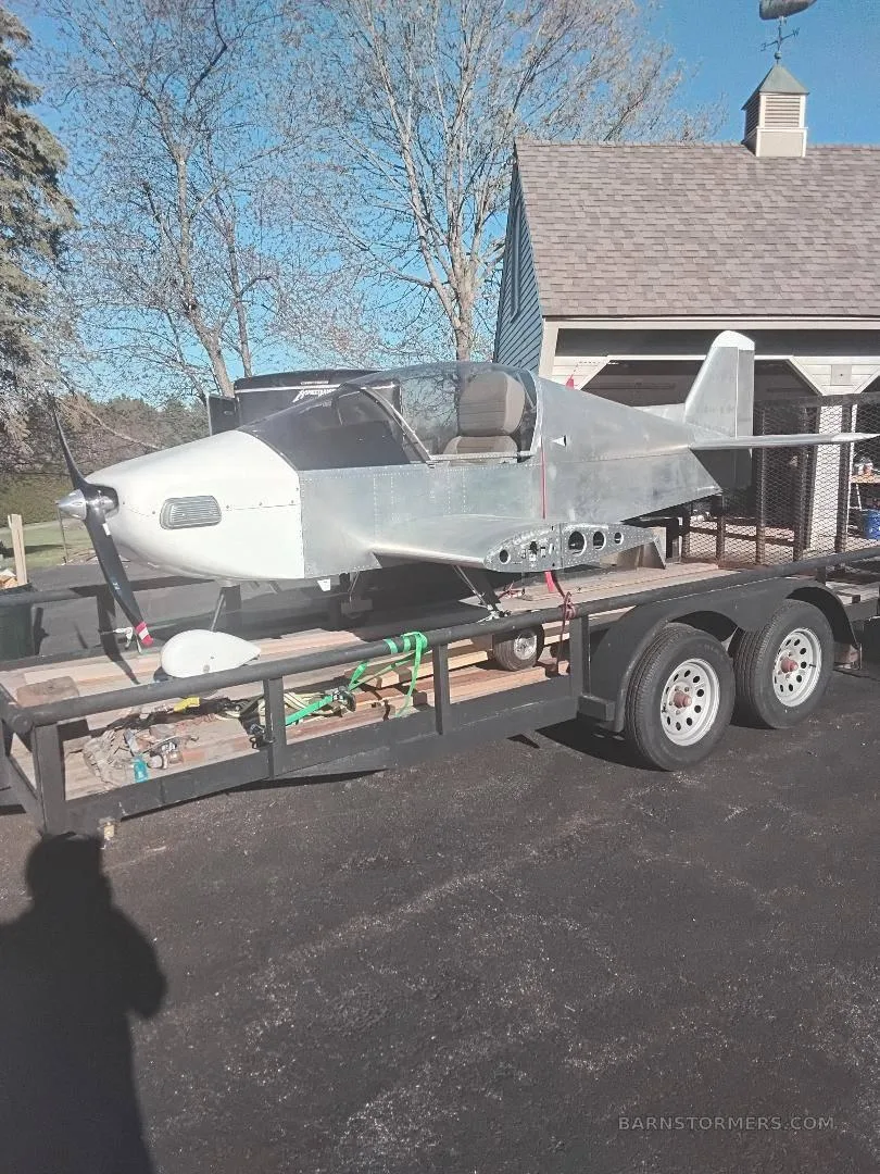An unpainted experimental aircraft with a white nose cone, secured on a black utility trailer in a driveway.