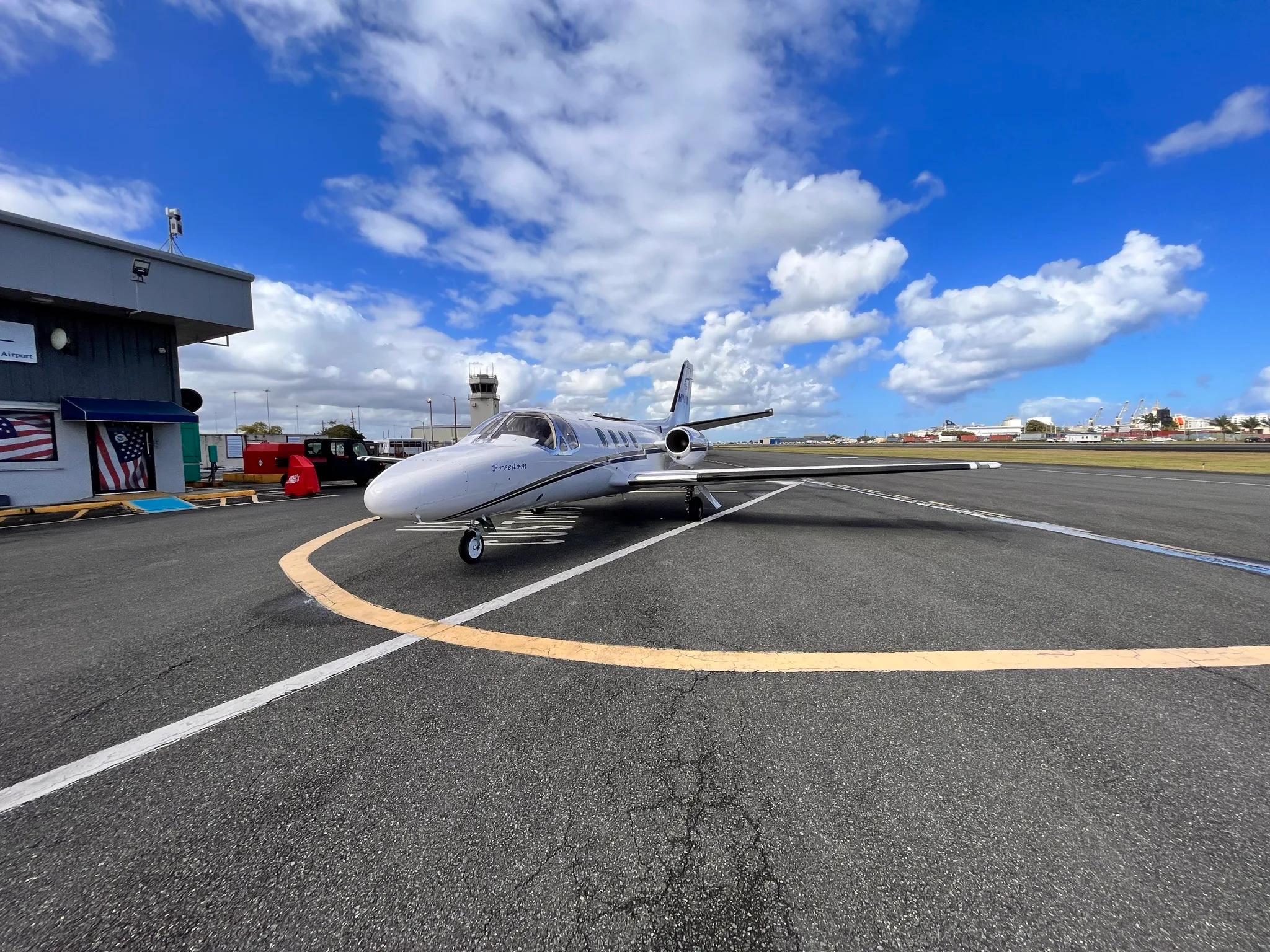 A white Cessna Citation Mustang with black and gold stripes sits on the tarmac in front of a building.