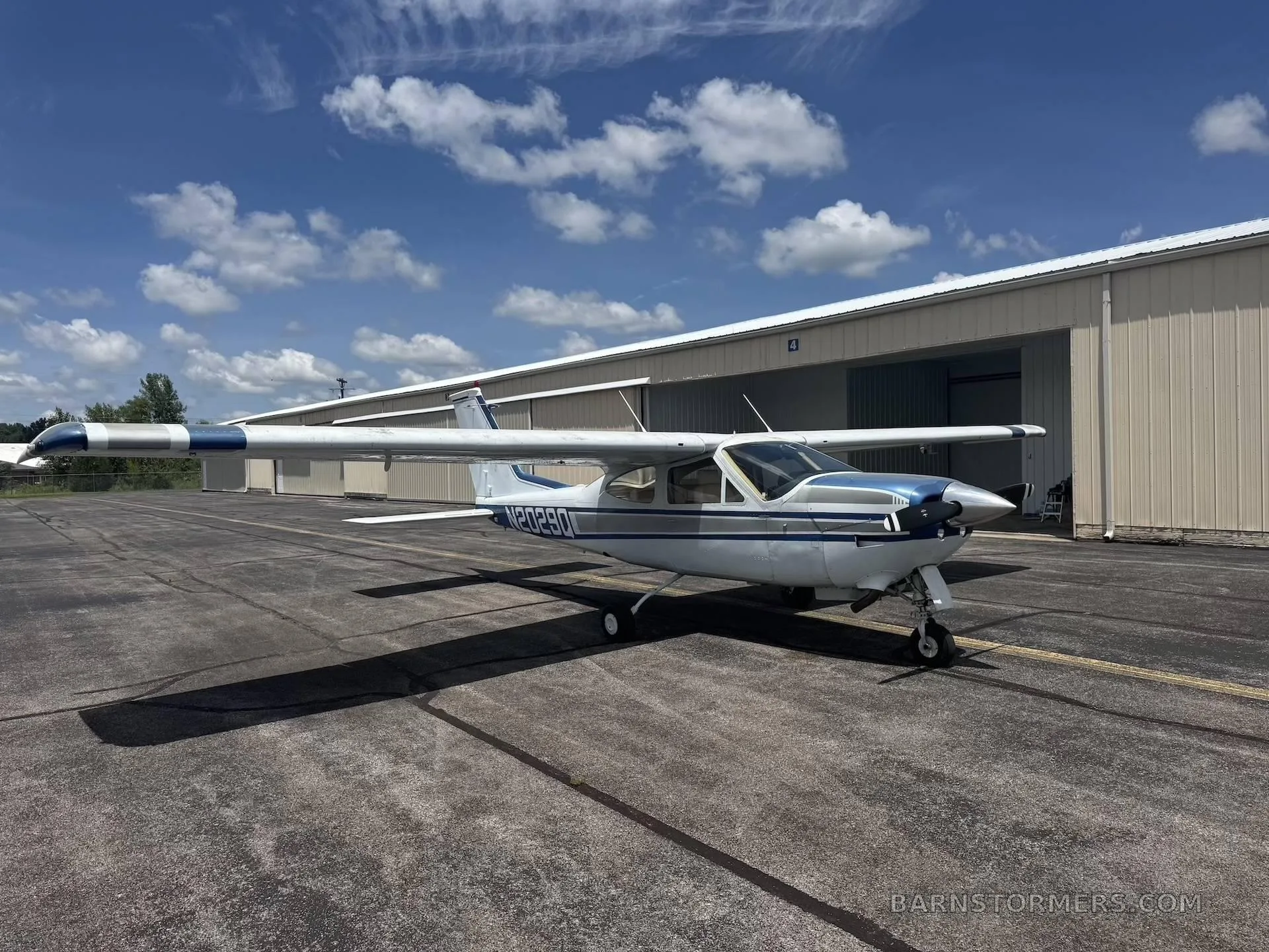 A white and blue Cessna aircraft, N20290, parked on a tarmac in front of hangars under a partly cloudy sky.