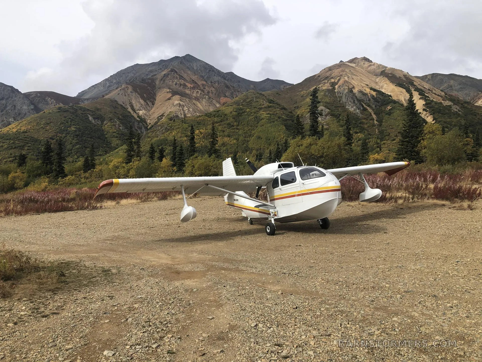 A white Republic RC-3 Seabee seaplane with red and yellow stripes parked on a gravel field with mountains in the background.