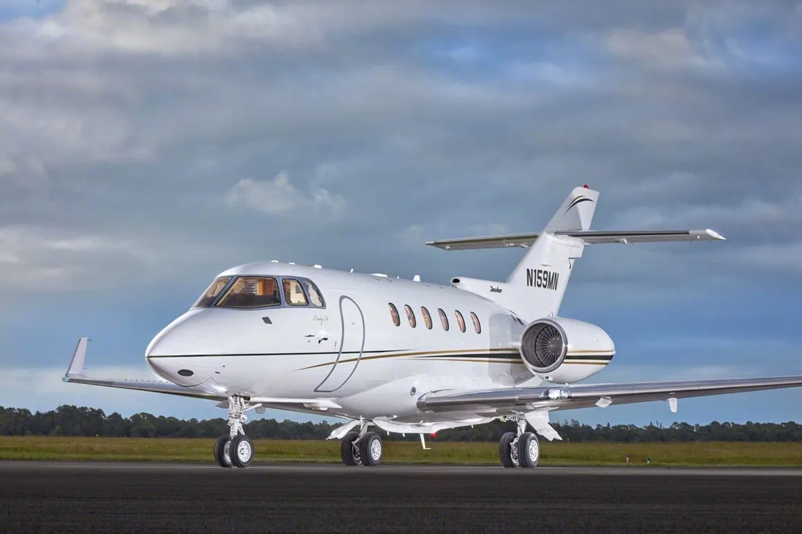 A white business jet with gold and black stripes, tail number N159MN, parked on a tarmac under a cloudy sky.