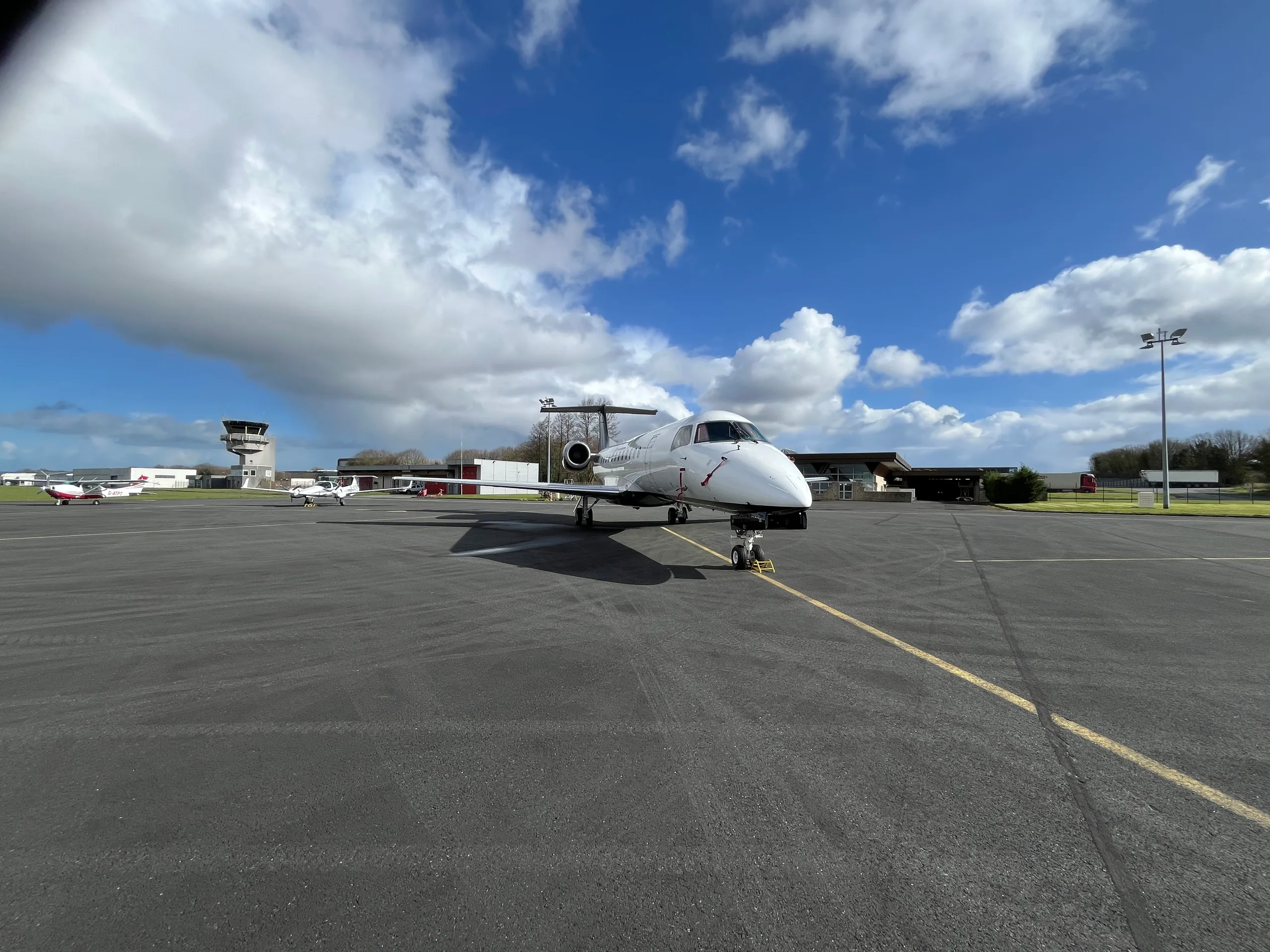 A white business jet with dark engines and tail, parked on an asphalt tarmac with a control tower in the background under a bright blue sky.