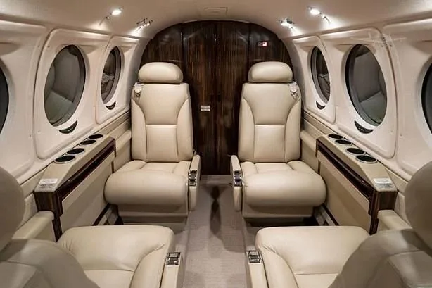 The aircraft cabin shows light beige leather seats in a club arrangement, with dark wood paneling on the rear bulkhead.