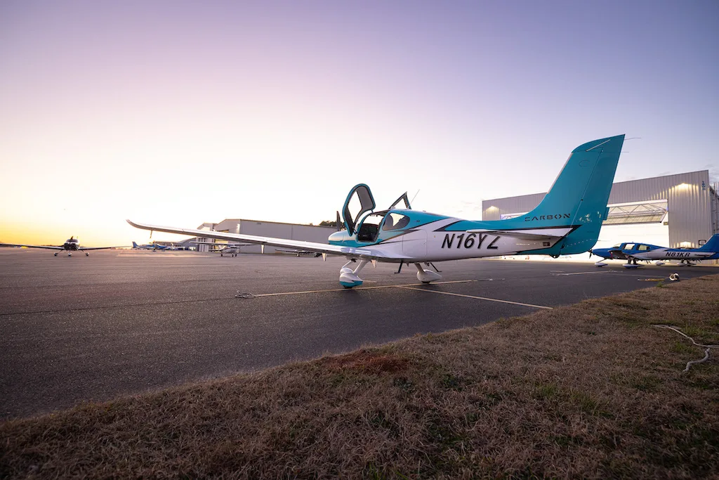 A turquoise and white Cirrus SR22 aircraft with its doors open, parked on an airfield next to hangars at sunset.