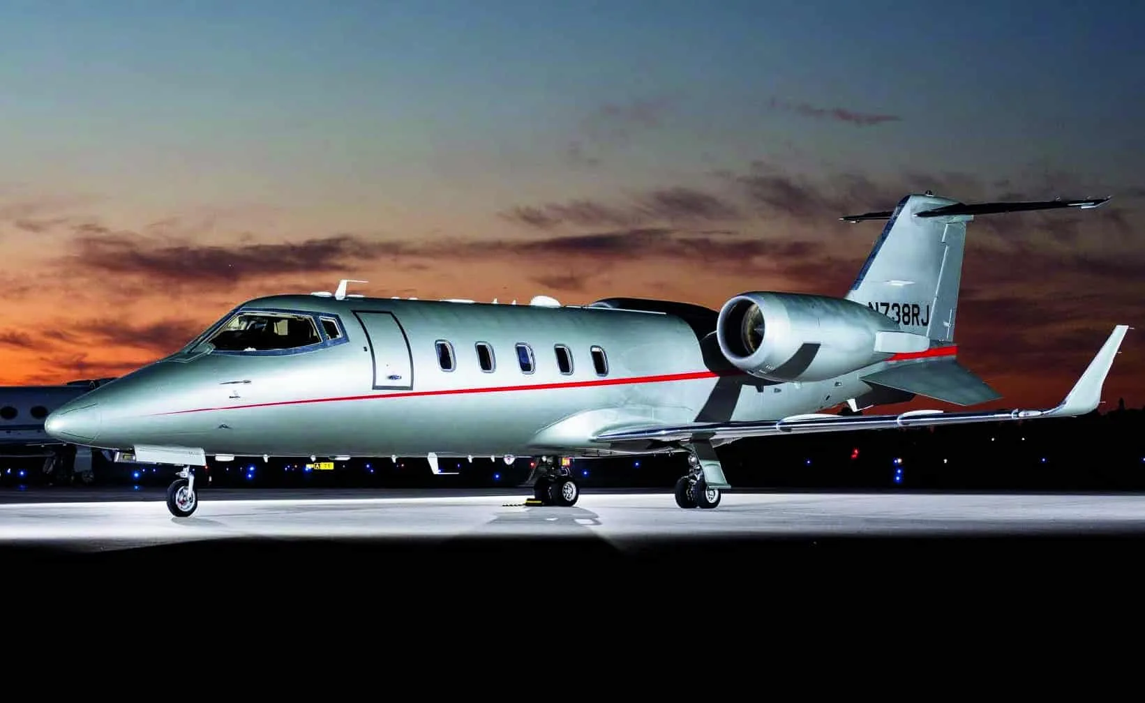 A silver Learjet 75, tail number N738RJ, with a red stripe, parked on a tarmac at sunset.