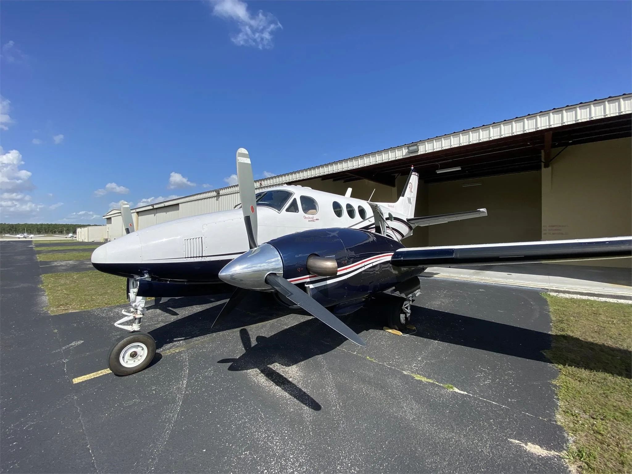 A front-quarter view of a white and dark blue Beechcraft King Air with propellers, parked on an asphalt apron.