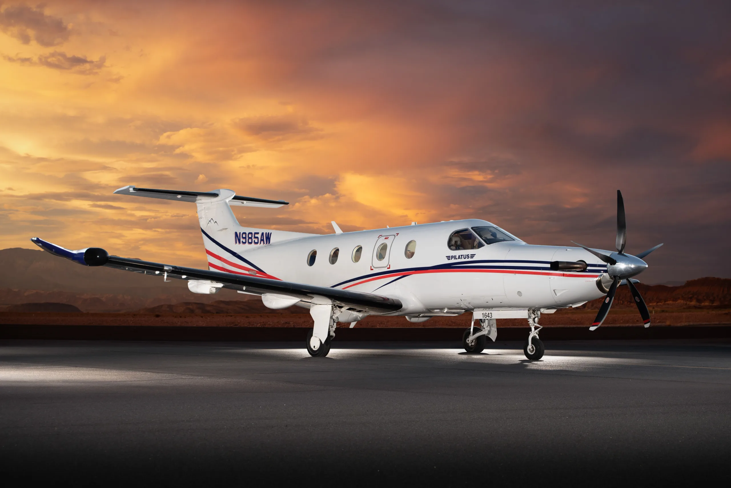 A Pilatus PC-12 aircraft, N985AW, with a white, blue, and red paint scheme, sits on a tarmac at sunset from a slightly different angle.