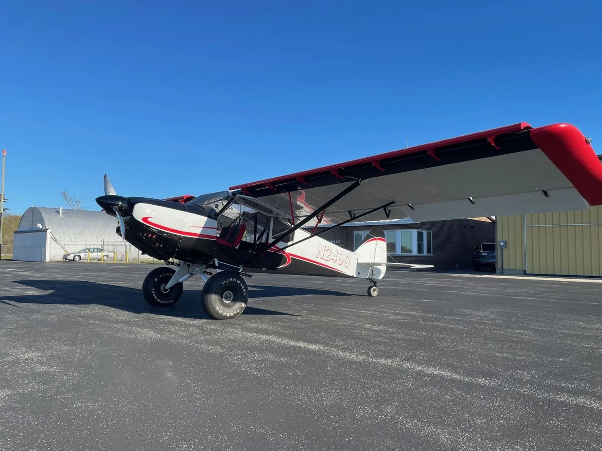 A white, black, and red STOL Quest 4 aircraft (N24SQ) is parked on an asphalt tarmac with hangars in the background.