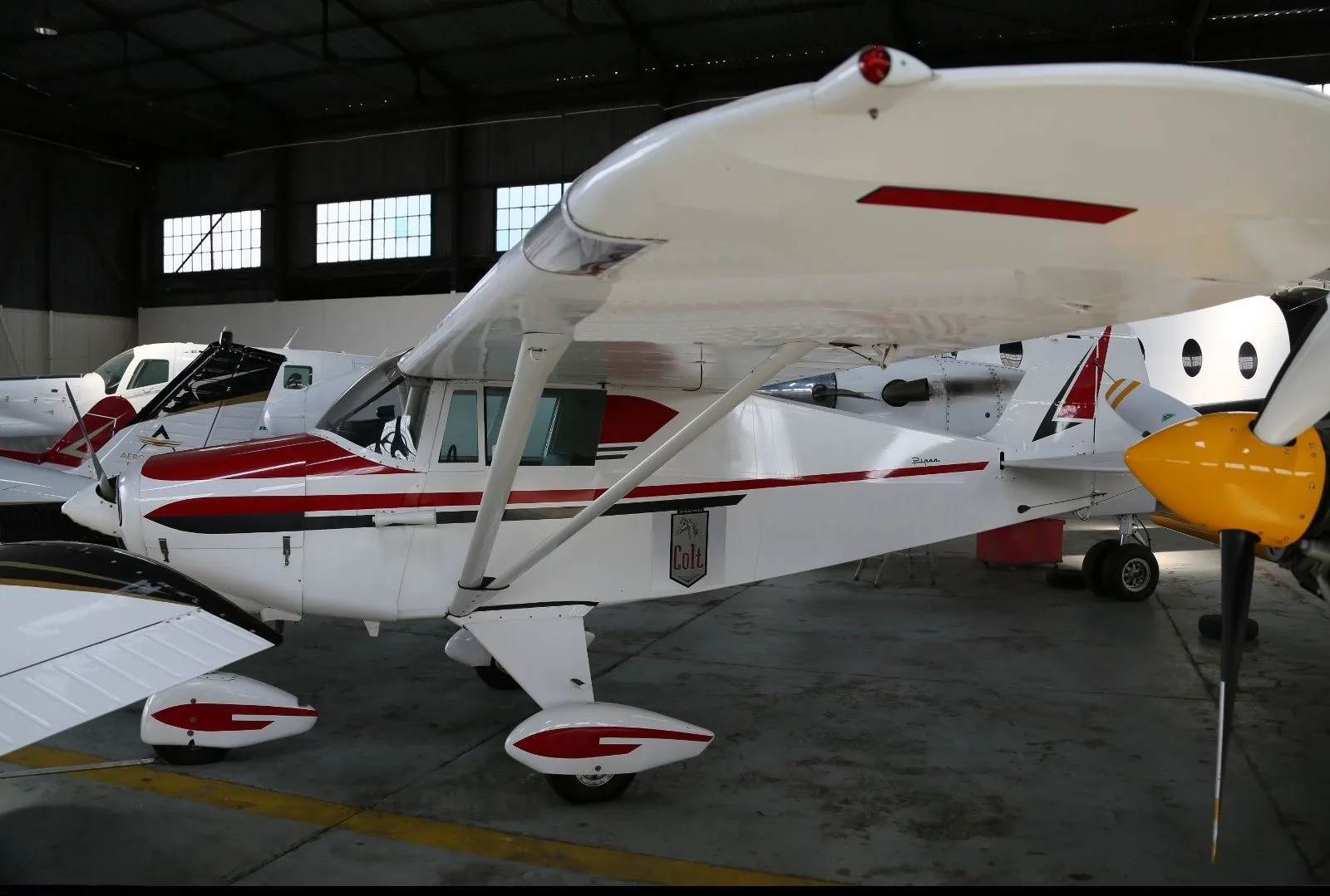 A white Piper Colt high-wing aircraft with red stripes and a yellow propeller tip, parked in a hangar.