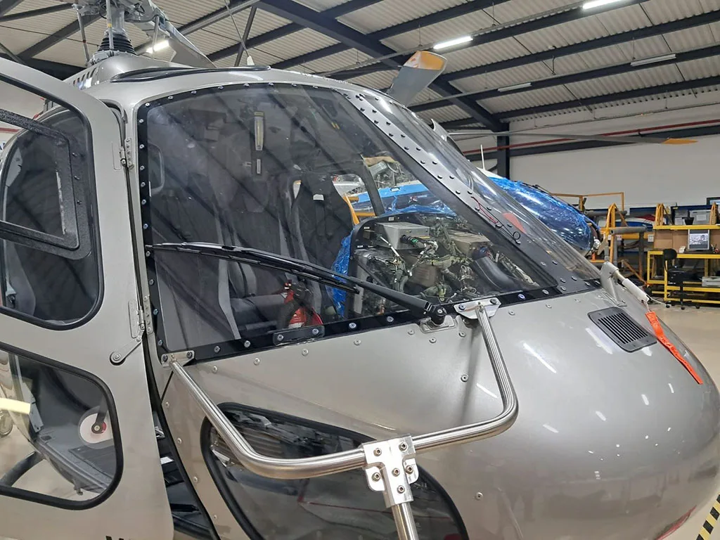 A close-up view of the front windshield and nose of a silver Airbus H125 helicopter inside a hangar.