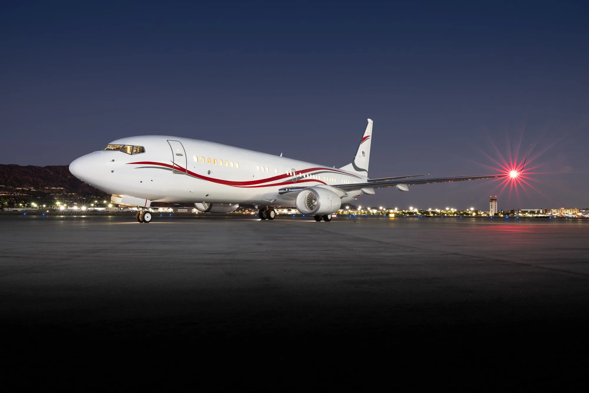 A Boeing BBJ aircraft with a white and red livery parked on a tarmac at dusk, with city lights in the background.
