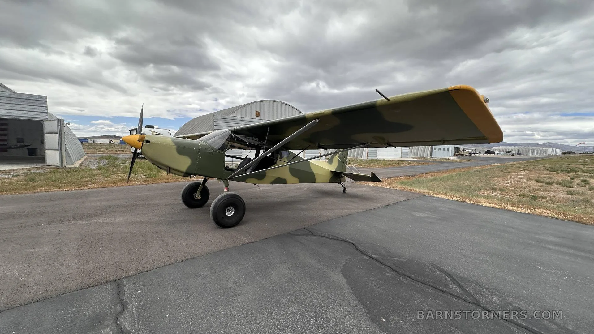 A light aircraft with camouflage paint and a yellow propeller spinner is parked on an asphalt runway with hangars in the background.