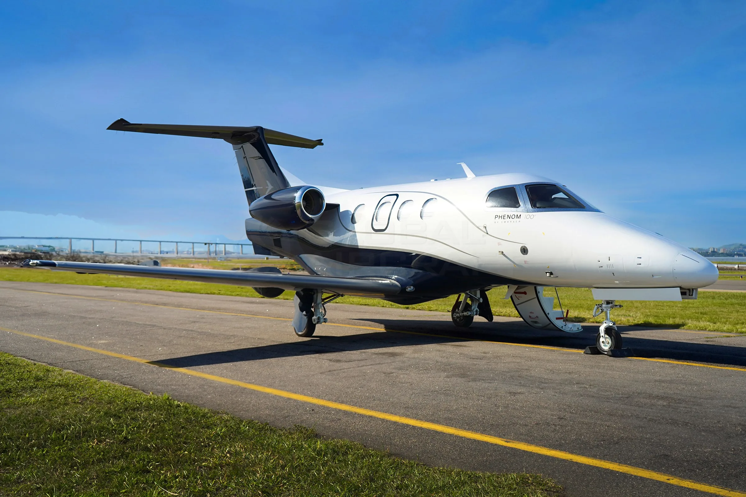 A white and black Embraer Phenom 100 business jet is parked on an asphalt runway with green grass and a bridge in the background.