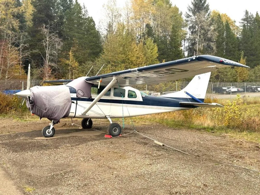 A white and blue Cessna 185 aircraft with a grey engine cover, parked on a gravel patch with trees in the background.