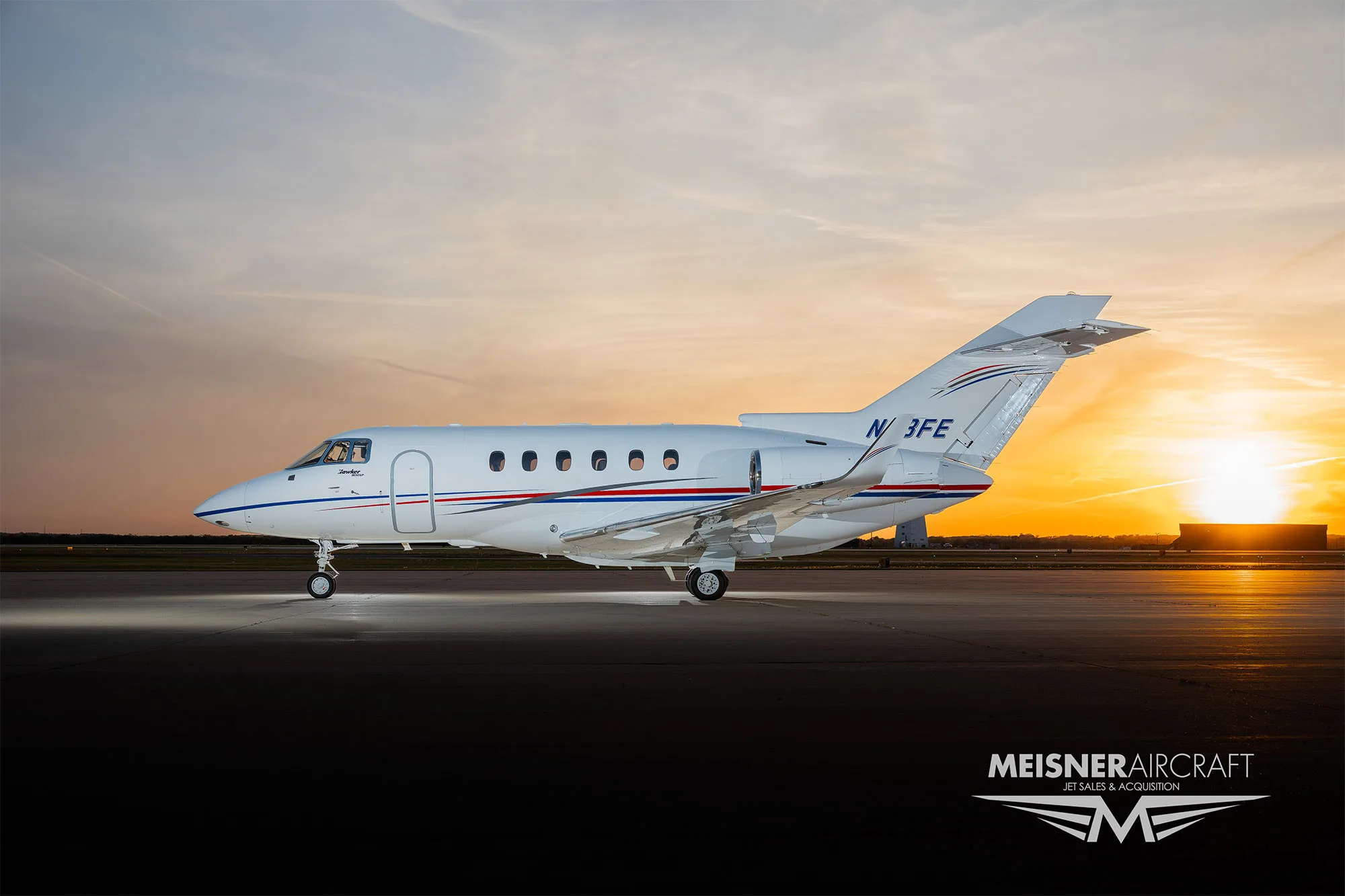 A white business jet with red and blue stripes, registration N13FE, parked on an airfield at sunset, viewed from the side.