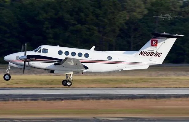 A white Beechcraft King Air 350 with black and red stripes, registration N208BC, is seen on a runway with its landing gear extended.