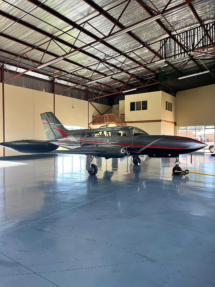 A dark grey twin-engine aircraft with red and silver accents, viewed from the left side in a hangar.