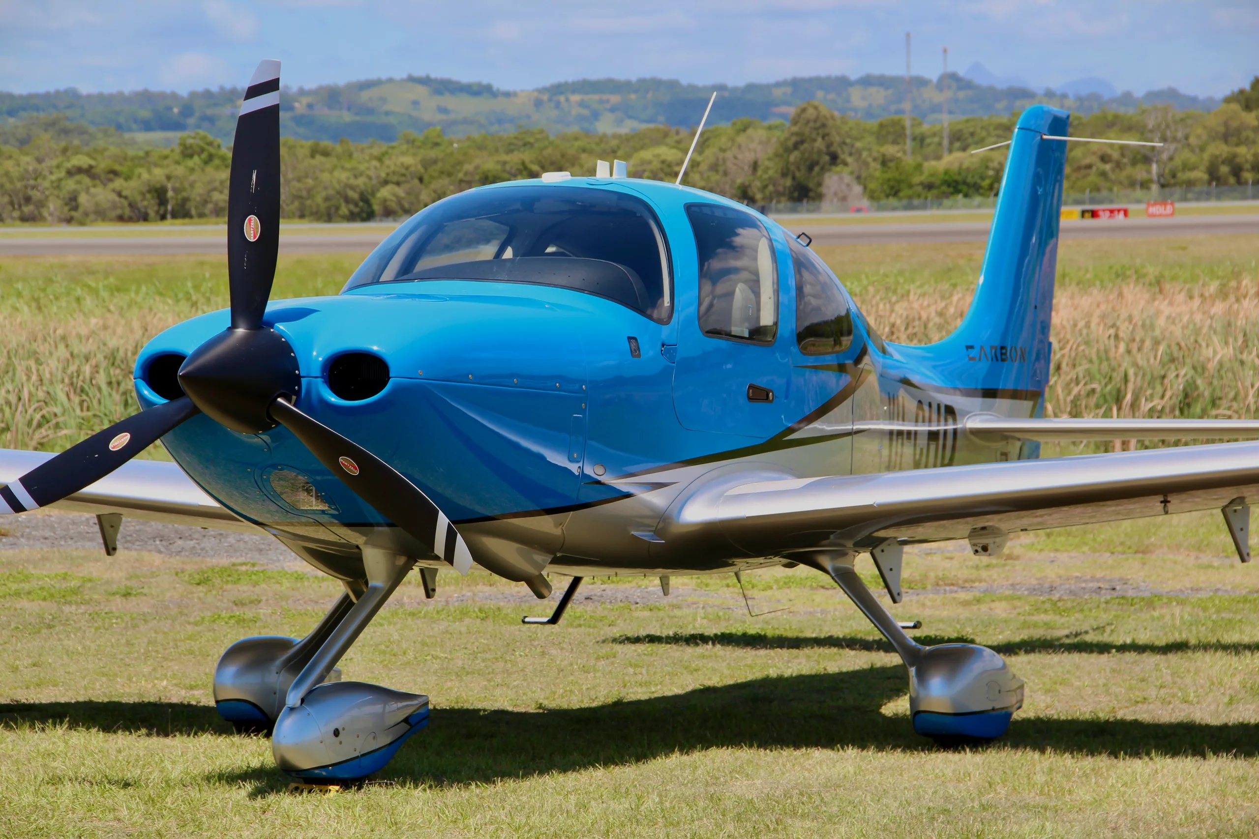 A blue and silver single-engine propeller aircraft with a three-bladed propeller is parked on a grassy field at an airport.