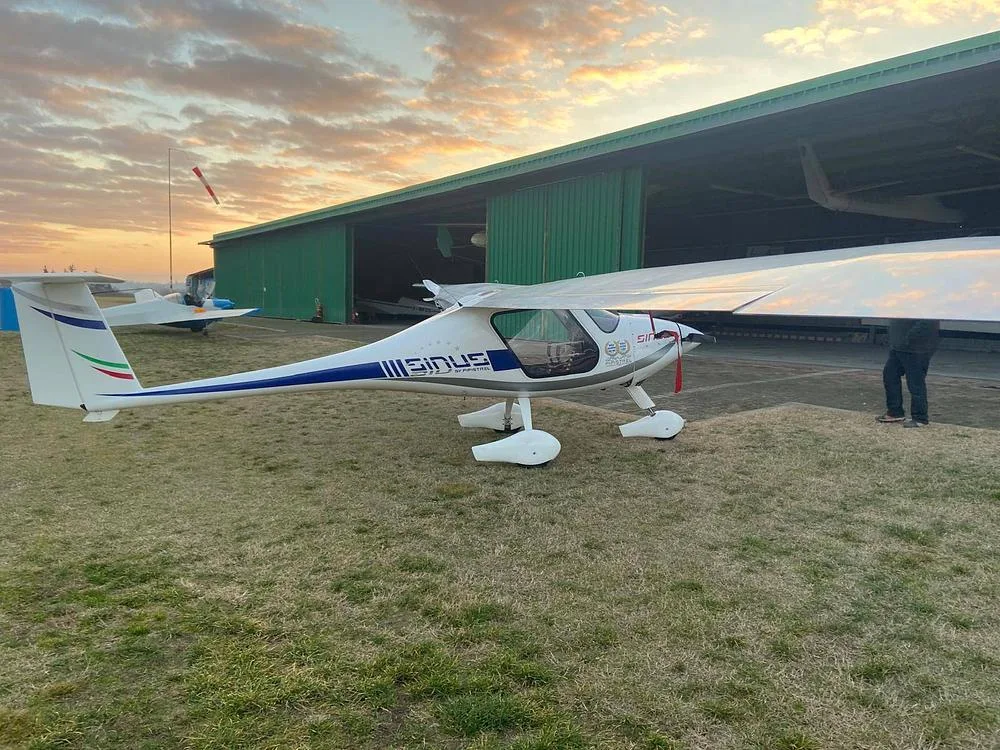 A white Pipistrel Sinus aircraft with blue and green tail stripes, parked on a grassy area in front of a green hangar at sunset.