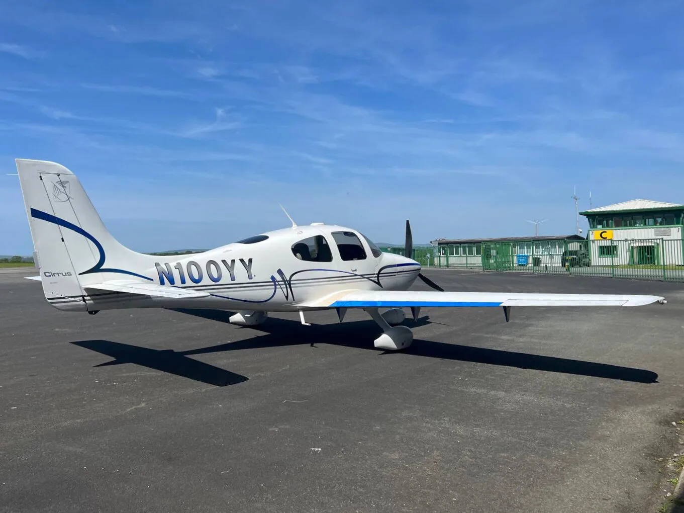 A white Cirrus SR22 aircraft with blue and grey stripes parked on an airfield tarmac under a blue sky.