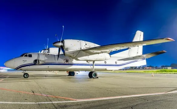 A white An-32 turboprop aircraft with blue stripes parked on a tarmac at night.