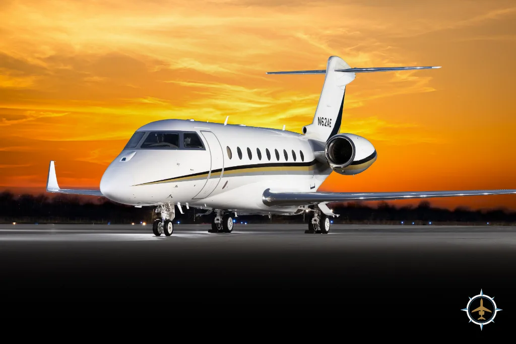 A white business jet with gold and black stripes on the tail, registration N62AE, parked on a tarmac at sunset.