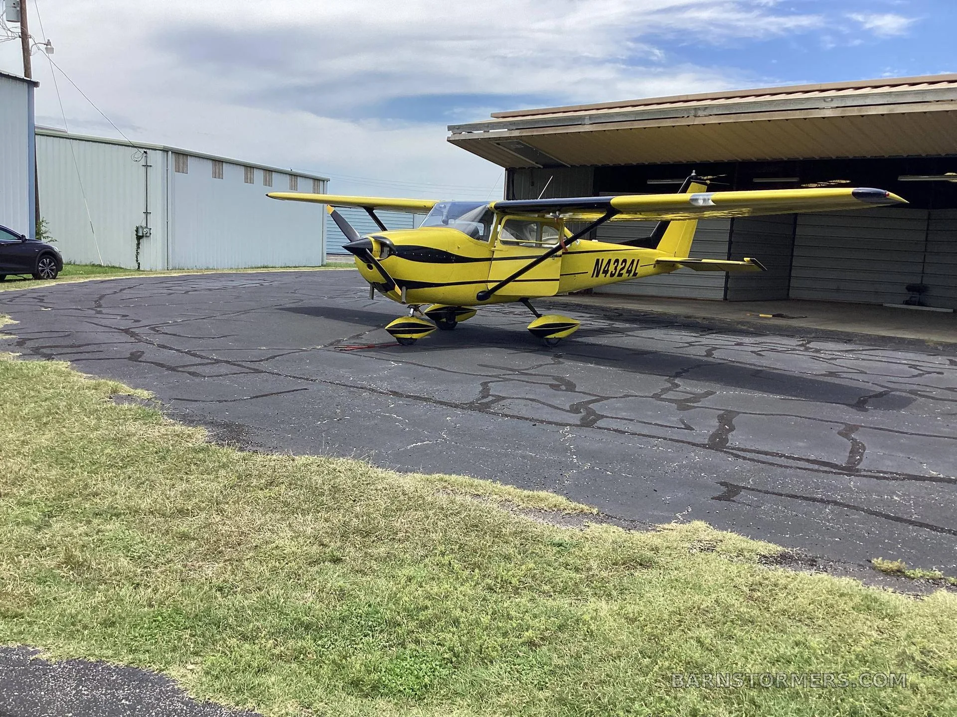 A yellow single-engine propeller aircraft with black stripes and registration N4324L is parked on an asphalt ramp next to a hangar under a cloudy sky.