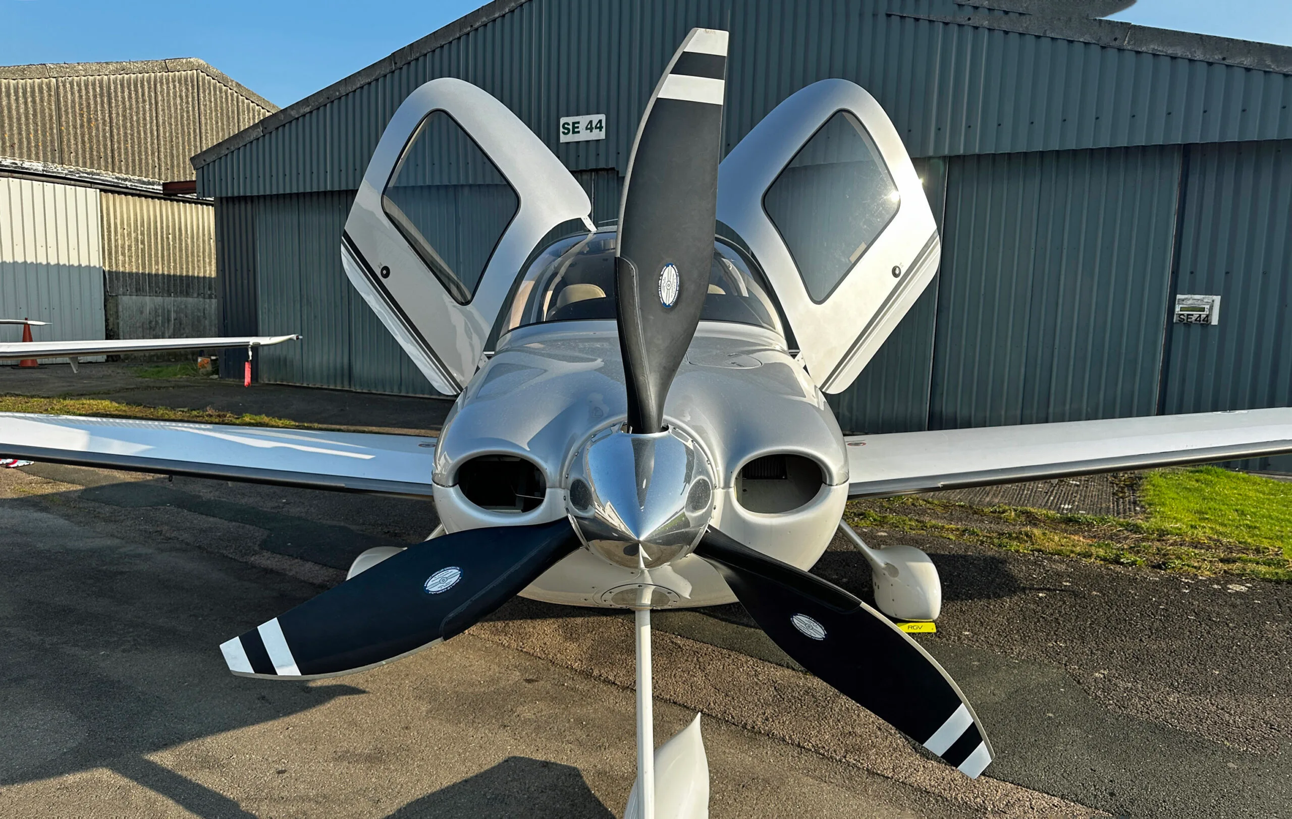 Front view of a silver and white Cirrus SR22 Turbo aircraft with open doors, parked in front of a hangar.